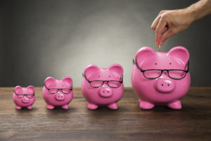Close-up Of Person Hand Inserting Coin In Pink Piggybank On Table