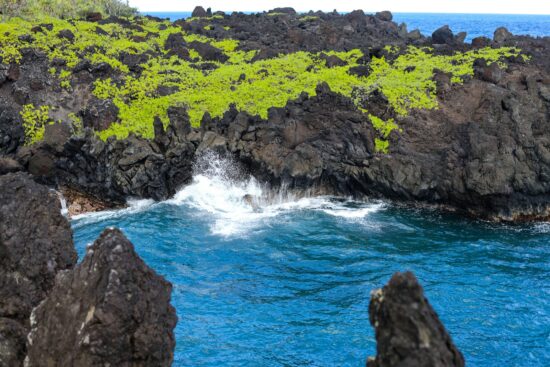 A striking coastal view in Hana, Maui, where the deep blue waters meet rugged volcanic rocks adorned with vibrant green foliage. The waves crashing against the shoreline create a mesmerizing contrast of colors and textures, showcasing the raw beauty of Hawaii’s untouched landscapes.