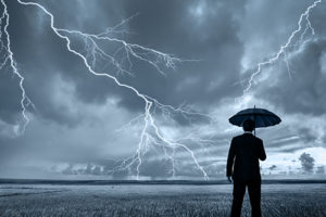 A rear view of a businessman in a suit, with an upheld umbrella, standing in a large field during a thunderstorm. Lightning is seen descending from a gray and cloudy sky.