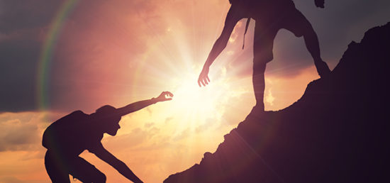 Man is giving helping hand. Silhouettes of people climbing on mountain at sunset.