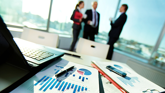 Business men and women meeting in background behind computer and data sheets.