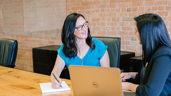 a dark-haired woman wearing glasses sits with another dark-haired woman at a wooden table in front of a notepad and laptop computer, possibly discussion fundraising options
