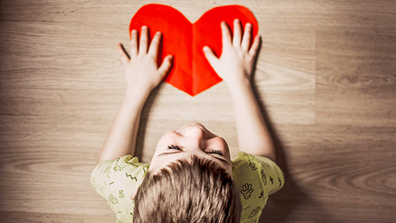 a young boy in a green shirt, holding both hands on a red heart on a wooden table; image used for a blog about tax savings of adopting a child