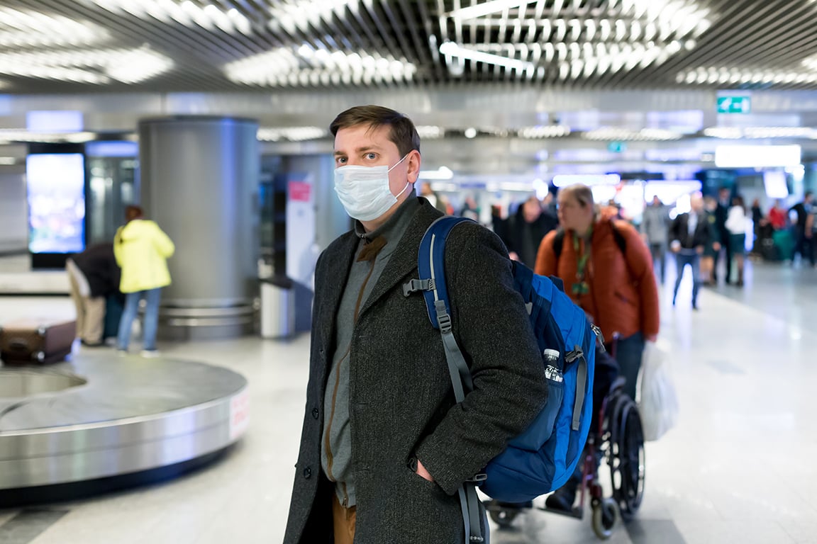 Young European man in gray coat, protective disposable medical mask in airport. Afraid of dangerous N-CoV 2019 influenza coronavirus mutated and spreading in China. Blue backpack, suitcase on wheels