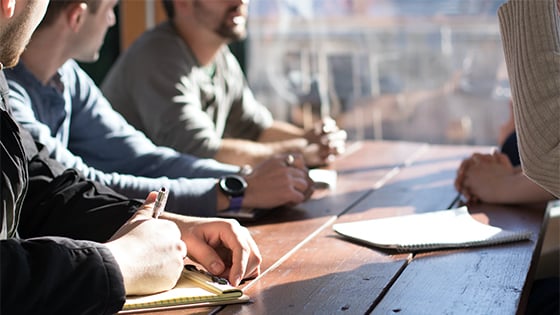 close up angle of three people sitting at a wooden table near a window overlooking the city with pens and notepads; image used for blog post for IRS new regulations for not-for-profits UBIT calculation