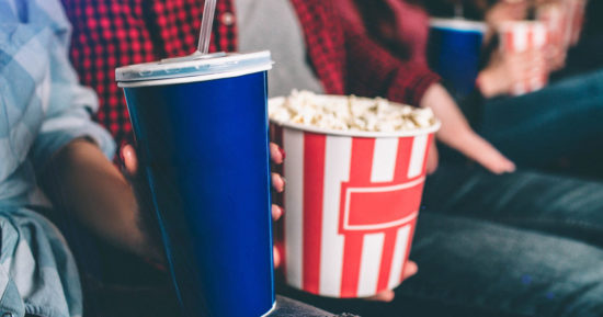 Close up of tasty but unhealthy food. There are basket of popcorn and a blue cup of coke on picture. Man and woman are holding it together.