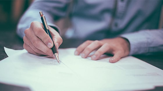 man in a blue button down shirt holds a pen to paper to sign documents; image used for blog post about new tax form 1099-nec