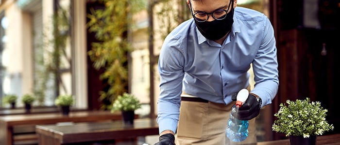 Waiter wearing protective face mask while disinfecting tables at outdoor cafe.;