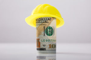 Close-up Of Yellow Hard Hat Over Rolled Up American Banknote On White Background