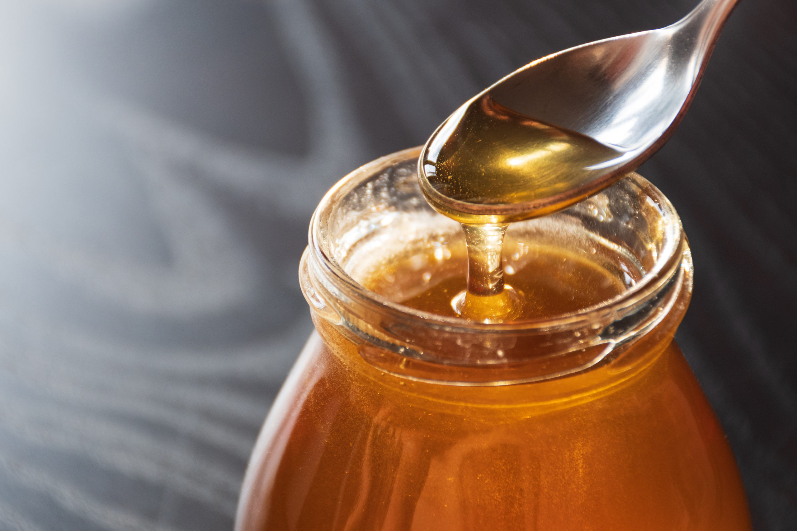 Delicious organic natural bio honey pours out of the spoon into a glass jar on wooden table with reflection of the sun, black background, close up