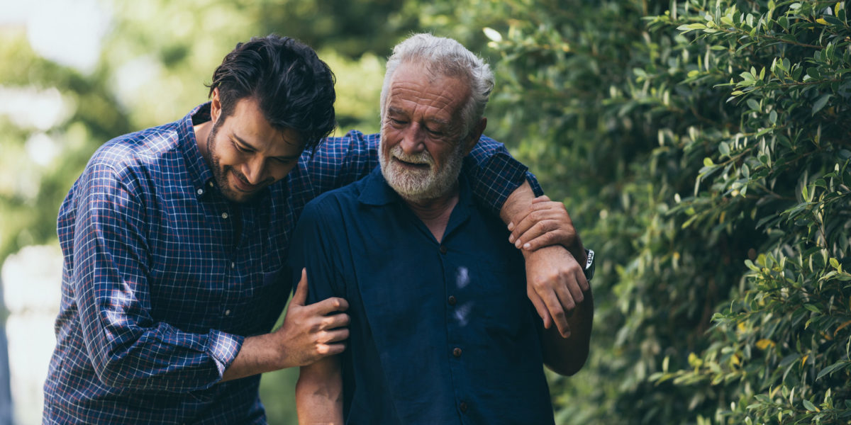 The old man and his son are walking in the park. A man hugs his elderly father. They are happy and smiling