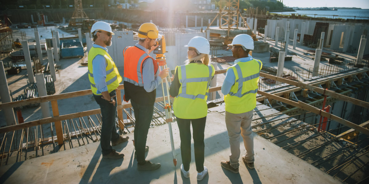 Construction Worker Using Theodolite Surveying Optical Instrument for Measuring Angles in Horizontal and Vertical Planes on Construction Site. Engineers and Architect Discuss Plans Next to Surveyor.
