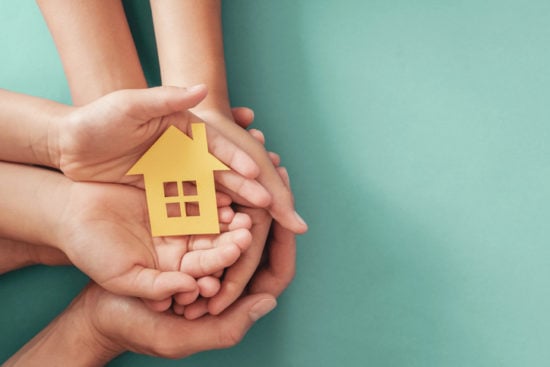 Hands holding yellow paper house on blue background, family home, homeless shelter housing and home protecting insurance, mortgage concept, foster home care