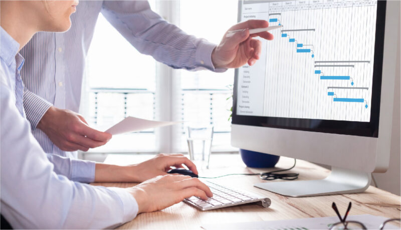 One coworker sits and types while another coworker stands by and points at the data on the computer screen