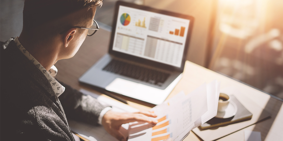 Young finance market analyst in eyeglasses working at sunny office on laptop while sitting at wooden table.Businessman analyze document in his hands.Graphs and diagramm on notebook screen.Blurred.