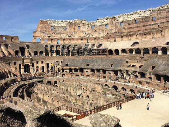A large stone structure with many round arches with Colosseum in the background Description automatically generated with medium confidence