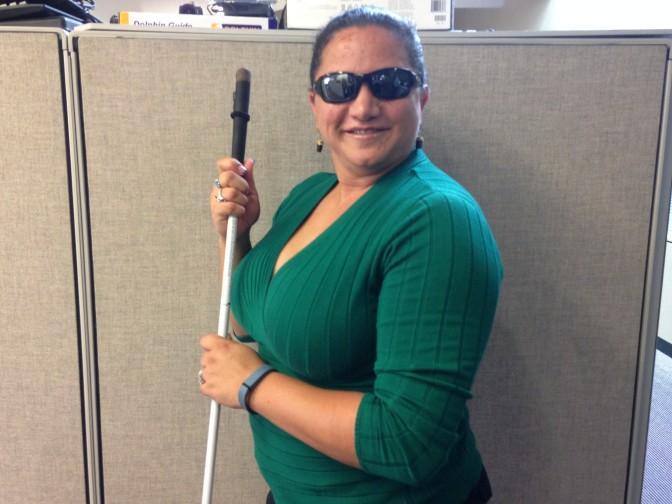 Lisamaria Martinez, wearing dark glasses and standing in an office, holds a white cane and is turned towards the camera and smiling.