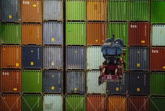 Close-up of multicolored stacked shipping containers with a container spreader from a crane positioned in front, ready for loading or unloading operations.