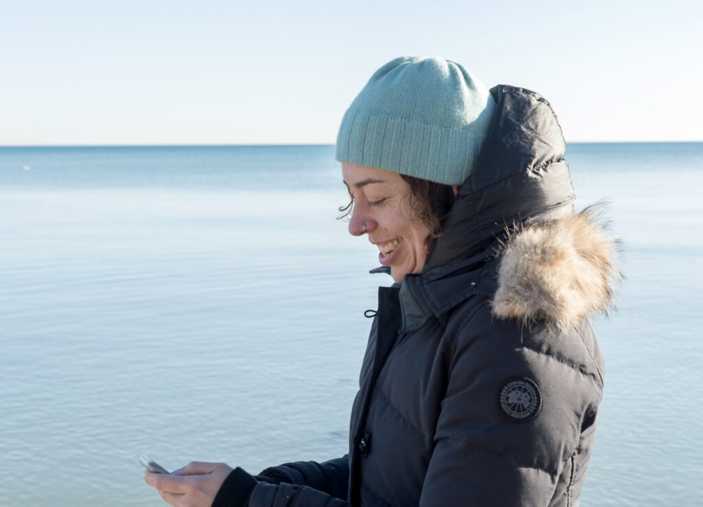 Woman in a hat and coat looking at her phone in front of a lake