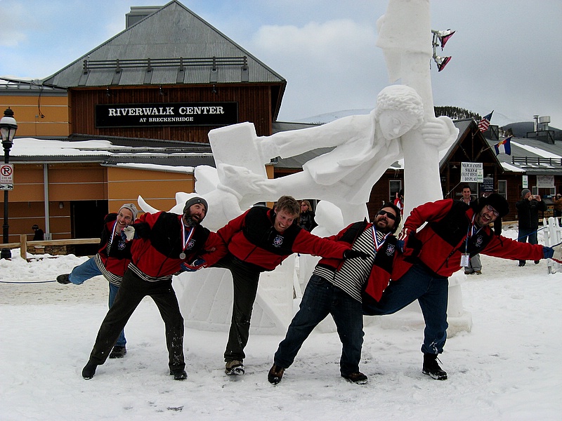 Finished snow sculpture of boy and dog hanging on to a light post against the wind with the artists stranding in front trying to "stay" upright against the wind.