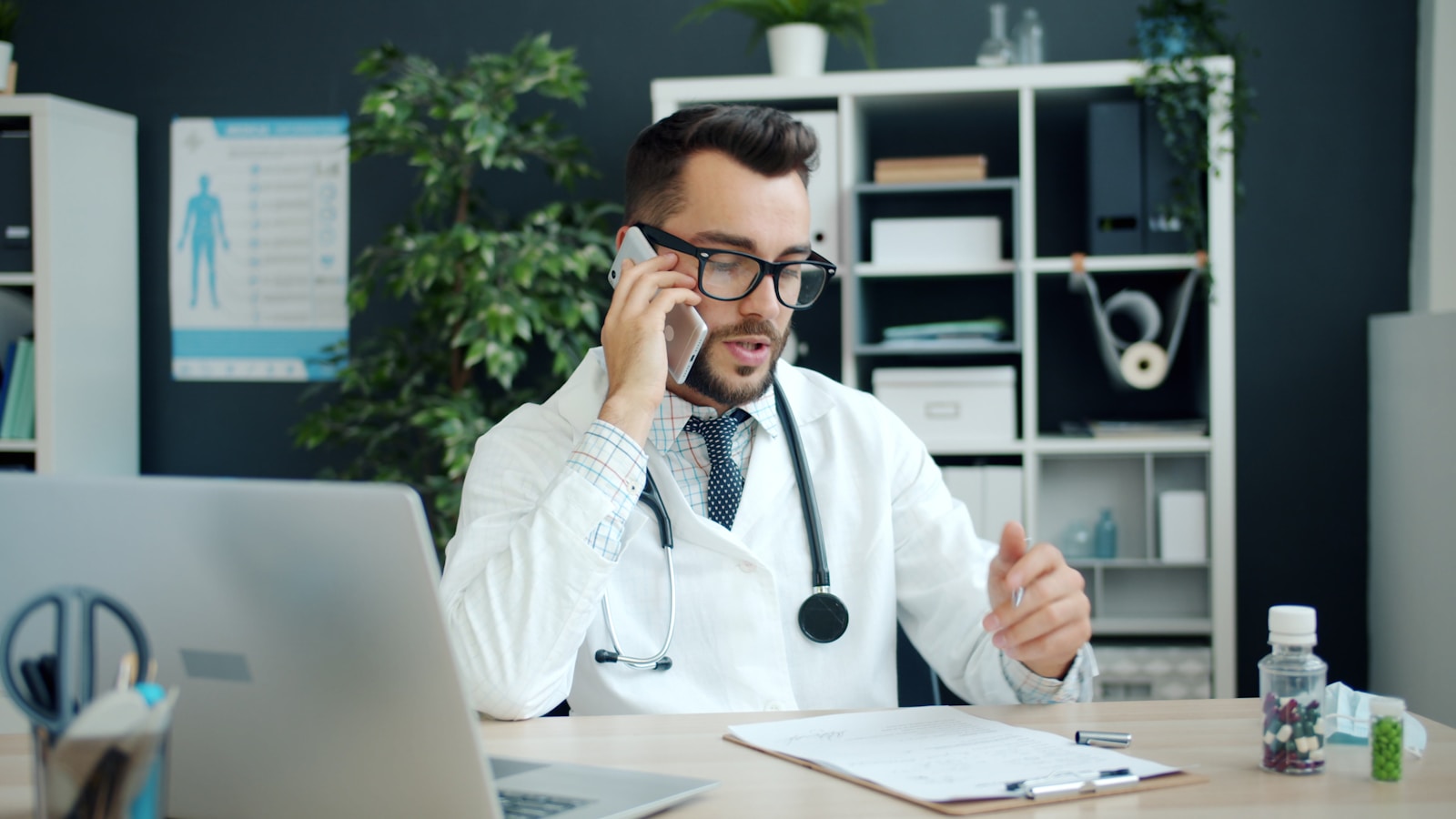 Young cheerful doctor is speaking on mobile phone, writing in chart and using laptop computer working in office alone. Modern devices, medicine and communication concept.