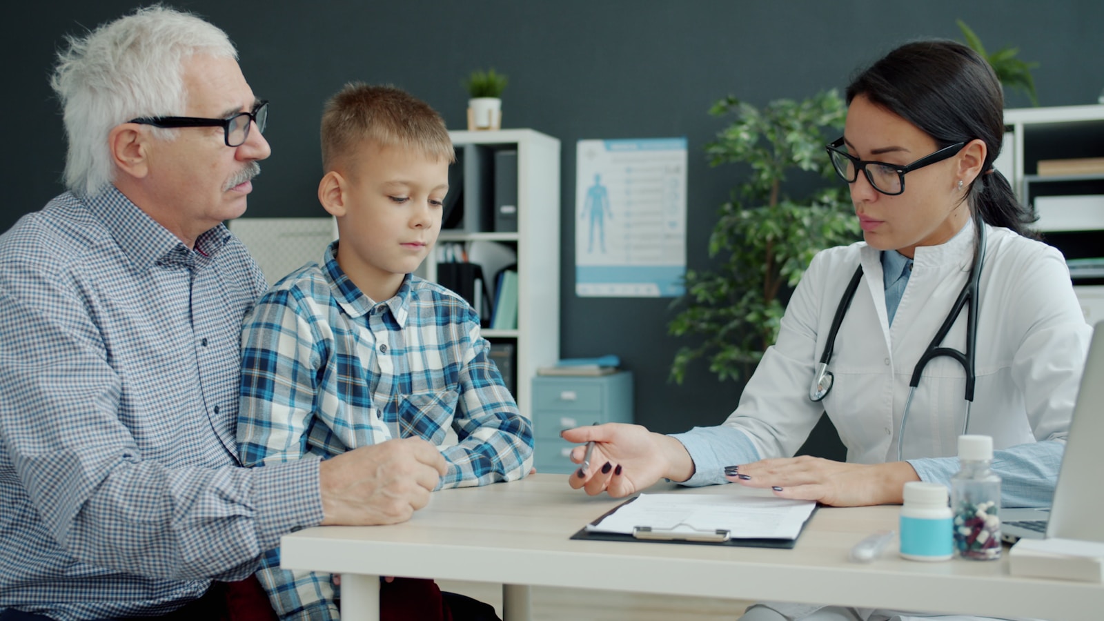 Female doctor is talking to caring grandfather and little grandson in office discussing child's health in hospital. Childhood, health and people concept.