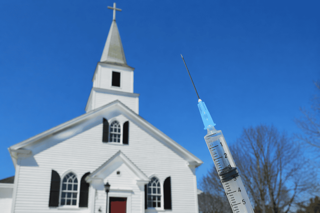 Ninth Circuit Denial of En Banc Review in Detwiler Reinforces the Tension in Religious Accommodation Cases 2 Photo of a traditional white chapel-style church with a steeple and cross in the background, slightly out of focus, set against a clear blue sky. In the foreground, a large, sharply focused vaccine syringe is angled upward, with visible measurement markings, creating a visual contrast between religious architecture and medical imagery.