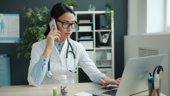 Young female doctor is talking on mobile phone and using laptop computer working in office alone. Modern devices, medicine and communication concept.