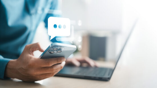 Person using smartphone beside laptop with chat icon overlay, representing online messaging, mobile communication, customer support, social media interaction, app notification, and digital connection.