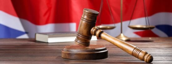 Judge's gavel, book and scales on wooden table against flag of United Kingdom-Shutterstock_2680090199