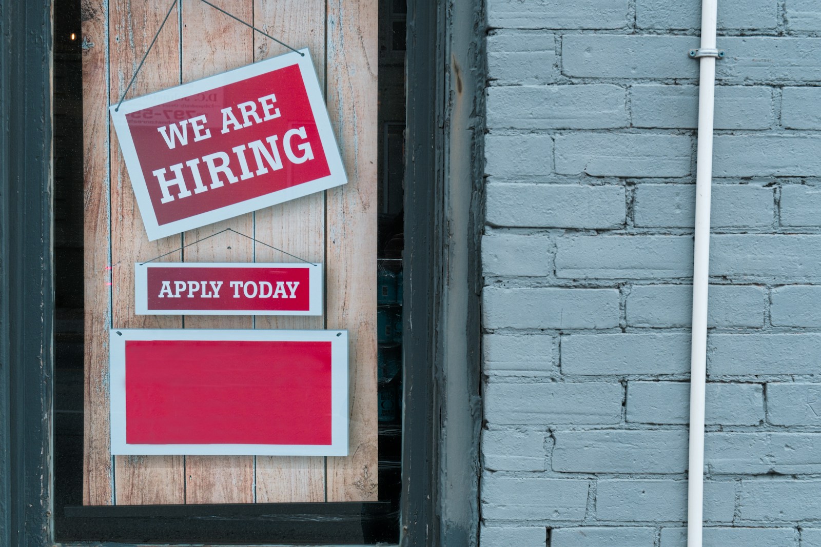 "We Are Hiring" Sign in Window of Brick Building - A red and white sign in a window of a brick building reads We Are Hiring with a smaller sign that says Apply Today.