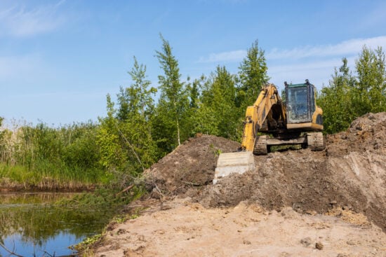 A yellow tracked excavator works along a wetland pond bank, moving soil and clearing vegetation for land development and erosion control. Bright blue sky and green trees frame the industrial scene