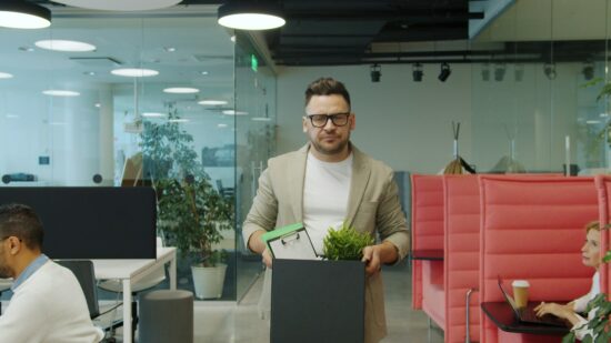 Fired middle-aged man in suit is walking in shared office leaving workplace with box of belongings feelings sad being unemployed. Dismissal and emotions concept.